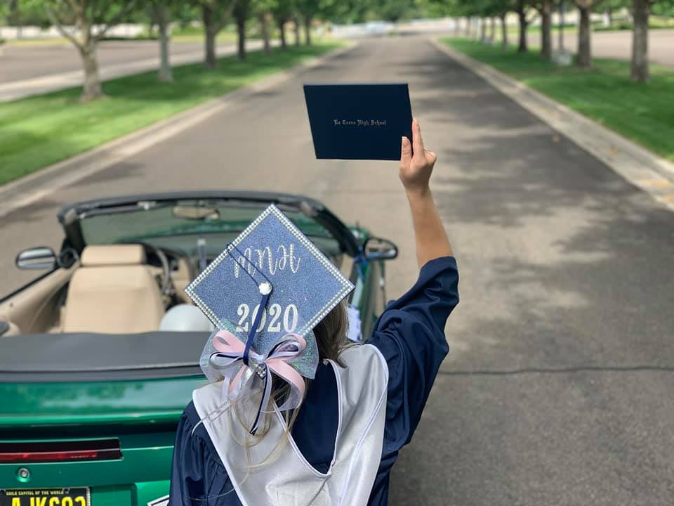 Girl wearing graduation cap and gown, standing next to car and holding up diploma